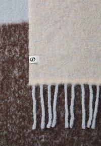 Couverture en laine beige, gris clair et marron avec une texture douce. Présente des franges et une petite étiquette avec un logo.