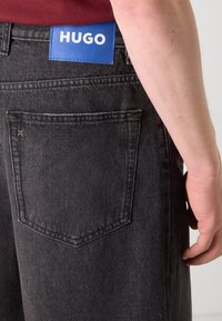 Gros plan d'une poche arrière de jean en denim noir avec une étiquette rectangulaire bleue portant la mention HUGO au-dessus de la poche, et une main d'une personne à côté de la poche.