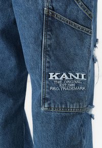 Blue denim jeans with a side pocket featuring white embroidered text "KANI THE ORIGINAL EST. 1989 REG. TRADEMARK" and slight distressing.
