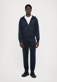 Ensemble de survêtement bleu marine comprenant une veste à fermeture éclair avec capuche et un pantalon ajusté assorti. Fabriqué en tissu lisse avec des poignets élastiques et des poches latérales.