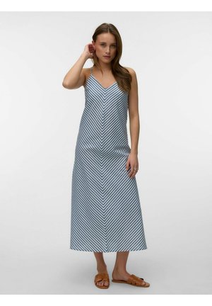 Femme en robe maxi sans manches à rayures chevrons bleues et blanches et sandales marron à enfiler, debout, une main touchant ses cheveux.