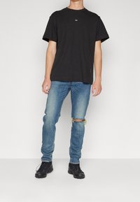 Svart bomullströja med rund hals och korta ärmar, kombinerad med slitna blå jeans med hål och texturerade svarta skor.