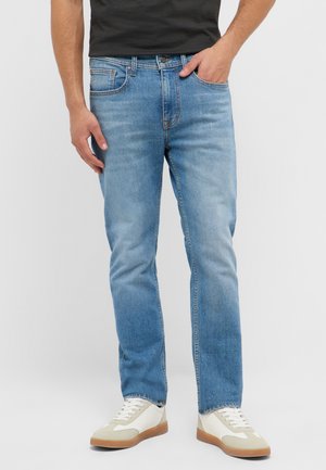 Mann trägt hellblaue gerade geschnittene Jeans, weiße Sneakers mit beigen Details und ein schwarzes Hemd, steht mit einer Hand in der Tasche.