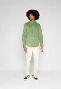Cămașă verde din catifea, cu nasturi și guler, purtată cu pantaloni crem și adidași alb-negru. Modelul stă în fața unui fundal gri.