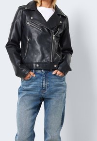 Czarna skórzana kurtka motocyklowa ze srebrnymi zamkami błyskawicznymi, zatrzaskami i usztywnionym kołnierzem, zestawiona z jasnoniebieskimi jeansami.