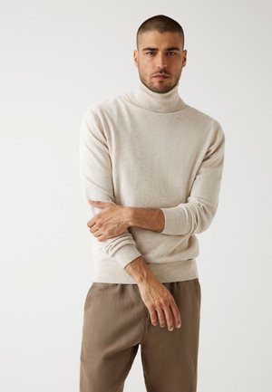Suéter de cuello alto beige en un material de punto suave, con un cuello y puños acanalados. Combinado con unos pantalones de corte relajado en color marrón claro.