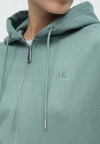 Ανοιχτό πράσινο φούτερ με φερμουάρ, κατασκευασμένο από απαλό ύφασμα, διαθέτει κουκούλα με κορδόνι και κεντημένο λογότυπο στο στήθος. Λείο υλικό και ασημένιο φερμουάρ.