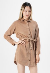 Vrouw met lang haar, gekleed in een tan kleurige ribfluwelen jurk met knopen, gestrikt in de taille, poseert met één hand op haar heup tegen een witte achtergrond.