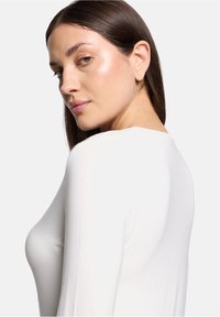 Weißes Langarmoberteil mit rundem Kragen, glatter Textur, figurbetontem Schnitt und leichtem Glanz, von der Seite betrachtet.
