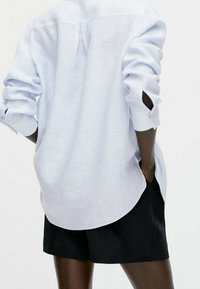 Lichtblauwe gestreepte blouse met opgerolde mouwen en een achterplooitje, gecombineerd met zwarte shorts, wat een relaxte pasvorm en een gestructureerde stof laat zien.