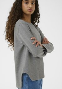 Giovane donna con lunghi capelli ricci che indossa un top grigio a maniche lunghe e jeans blu, in piedi con le braccia incrociate su uno sfondo bianco.