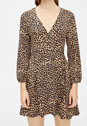 Kobieta w krótkiej sukience z długim rękawem i dekoltem w serek, z wzorem w cętki i dopasowaną talią.