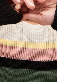 Pull en tricot avec des rayures horizontales vertes, noires, roses, jaunes et crème. Col côtelé et tissu texturé. Mains ajustant le pull.