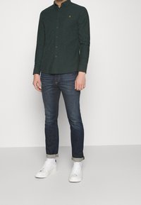 Camisa de color verde oscuro con botones y cuello, combinada con vaqueros azules y zapatillas blancas. La camisa tiene un logo bordado en el pecho.