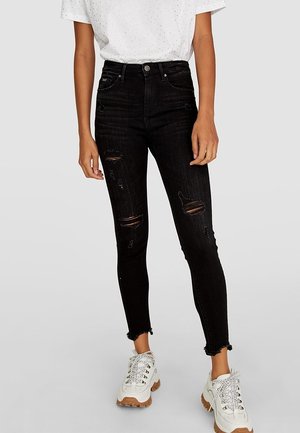 Personne portant un jean skinny noir déchiré taille haute avec des ourlets effilochés et des baskets blanches épaisses sur un fond uni.