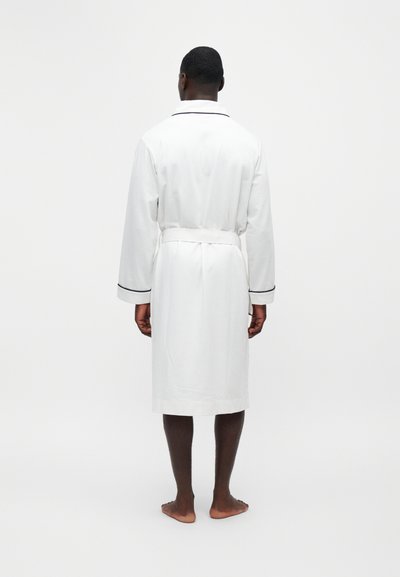 Homme debout pieds nus portant une robe blanche mi-longue aux genoux avec une bordure noire et une ceinture nouée à la taille, de dos sur un fond blanc uni.