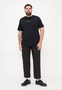 Black short-sleeve t-shirt with white text on the front, paired with dark brown pants and black shoes, standing on a white background.