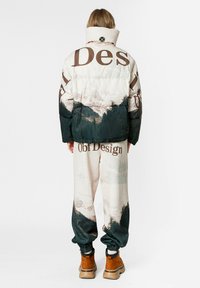 Chaqueta acolchada multicolor con texto marrón e imágenes de bosque, combinada con pantalones a juego que presentan el mismo diseño. Botas marrones completan el conjunto.