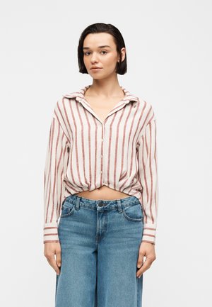 Γυναίκα με κοντά σκούρα μαλλιά, που φοράει λευκό πουκάμισο με κάθετες κόκκινες λωρίδες και μπλε τζιν ψηλόμεσα με φαρδιά πόδια, στέκεται μπροστά σε ένα απλό φόντο.