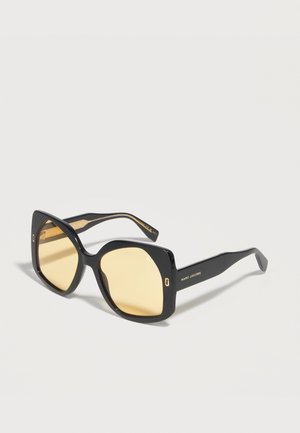 Store firkantede solbriller i sort med lysegule linser og Marc Jacobs-logo på siderne, vist mod en ensfarvet hvid baggrund.