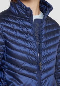 Navy insulated jacket with horizontal quilted design, high collar, full zipper, and branded detail on the inner placket. Smooth, glossy texture.