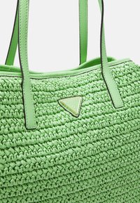 Světle zelená tkaná taška přes rameno s dvojitými popruhy a trojúhelníkovou nášivkou loga Guess ve středu přední strany.