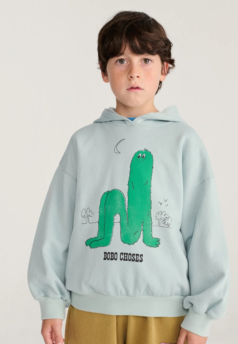 Garçon aux cheveux foncés portant un sweat à capuche bleu clair avec une créature verte de dessin animé et le texte "BOBO CHOSES" sur un fond uni.