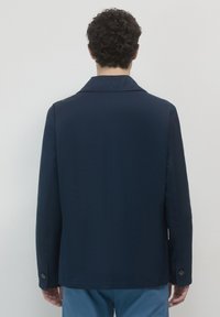 Uomo dai capelli ricci e scuri, che indossa un blazer blu navy e pantaloni azzurri chiari, in piedi di fronte a un muro grigio chiaro uniforme.