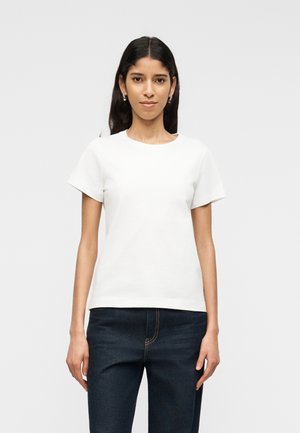 Hosszú sötét hajú nő ezüst karika fülbevalóval, egyszínű fehér rövid ujjú pólóban és sötétkék farmerben, fehér háttér előtt állva.