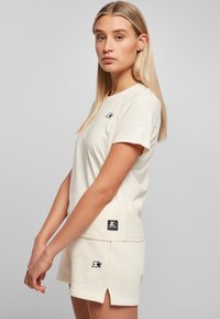 Kermavan vaate T-paita lyhyillä hihoilla, jossa on pieni musta logo rinnassa ja musta lappu helman kohdalla; yhdistettyinä matching-shortsien kanssa.