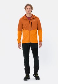 Veste légère bicolore orange et jaune avec capuche, fermeture éclair à l'avant, et pantalon noir avec accents texturés.