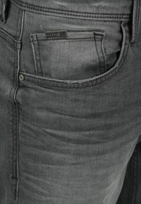 Jeans de denim gris que presentan un bolsillo frontal con cierre de botón de metal y etiqueta de marca. La tela tiene una ligera textura y un acabado desgastado.