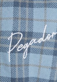 Blue and light blue checkered fabric with a textured weave, featuring white embroidered "Pegador" text in a cursive font.