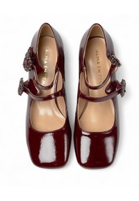 Scarpe Mary Jane in pelle verniciata bordeaux con punta quadrata, cinturini alla caviglia e dettagli con fibbie ornate. Finitura liscia e lucida con leggera imbottitura.