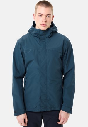 Junger Mann in einer dunkelblauen wasserfesten Jacke mit Kapuze, stehend vor einem schlichten hellen Hintergrund.