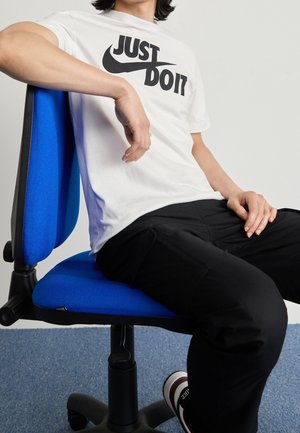 Camiseta blanca con texto negro "JUST DO IT", pantalones negros, sentado en una silla de oficina azul con un asiento texturizado, contra un fondo neutro.