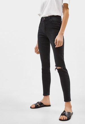 Person wearing black skinny jeans with a knee rip, white t-shirt, and black open-toe slide sandals standing on a plain white background.