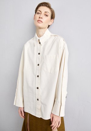 Κρέμ μακρυμάνικο πουκάμισο με κουμπιά και μία τσέπη στο στήθος, καφέ κουμπιά και άνετη εφαρμογή, σε στρώσεις πάνω από μια καφέ υφασμάτινη φούστα.