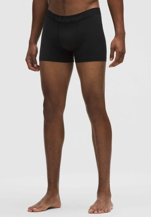 Uomo con boxer Lululemon neri, in piedi a piedi nudi contro uno sfondo semplice, gambe e parte bassa del torso visibili.