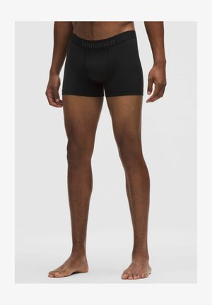 Mies, jolla on mustat Lululemonin bokserialusvaatteet, seisoo paljain jaloin yksiväristä taustaa vasten, näkyvissä ovat jalat ja alavartalo.