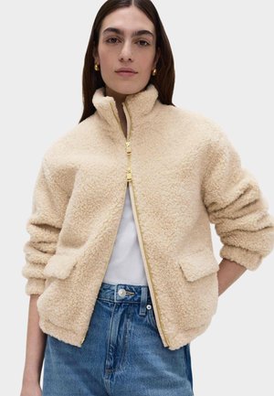 Beige fleecejack met een hoge kraag, ritssluiting en twee zakken aan de voorkant. Voorzien van geplooide mouwen en een zachte, textuurrijke stof.