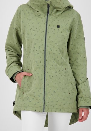 Veste à capuche verte avec petits motifs abstraits foncés, fermeture éclair à l'avant, poches latérales et manches longues, portée par une personne avec un pantalon blanc.