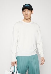 Weißer Strickpullover mit langen Ärmeln, rundem Ausschnitt und dezentem Branding; getragen mit türkisfarbenen Hosen und einer schwarzen Mütze mit gestickten Details.