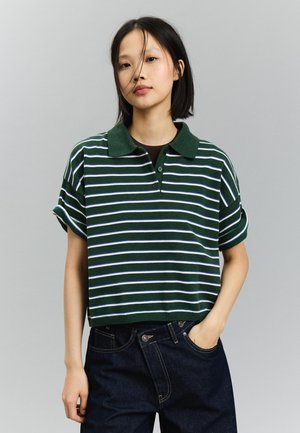 Junge Frau mit kurzen schwarzen Haaren, die ein grün-weiß gestreiftes Polohemd mit kurzen Ärmeln und dunkle Jeans trägt, mit einer Hand in der Tasche.