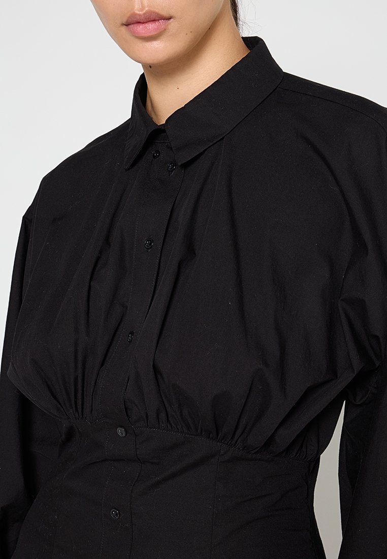 Frau trägt ein schwarzes Hemd mit Knöpfen, das unter der Brust gerafft ist, und einen klassischen Kragen vor einem schlichten Hintergrund.