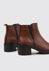 Botas de tobillo de piel marrón con tacón cuadrado, que presentan Cremalleras laterales y tachuelas plateadas a lo largo de la costura, textura suave y paneles elásticos.