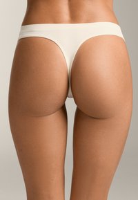 Beige thong, jossa on sileä ja joustava kangas. Siinä on leveä vyötärönauha ja minimaalinen peittävyys, mikä korostaa saumatonta muotoilua ja pehmeää tekstuuria.