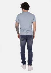 T-shirt in cotone azzurro chiaro con collo a giro, abbinato a jeans in denim blu scuro. L'outfit è completato da sneakers bianche e un tatuaggio sul braccio sinistro.
