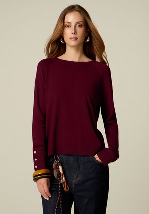 Frau trägt einen dunkelroten Langarmpullover mit Knopfdetails an den Bündchen, dunkle Jeans, große Creolen und mehrere Armreifen am linken Handgelenk.