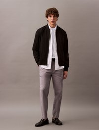 Black bomber jacket over a white shirt, paired with light grey trousers and dark leather shoes. Smooth textures and tailored fit.
